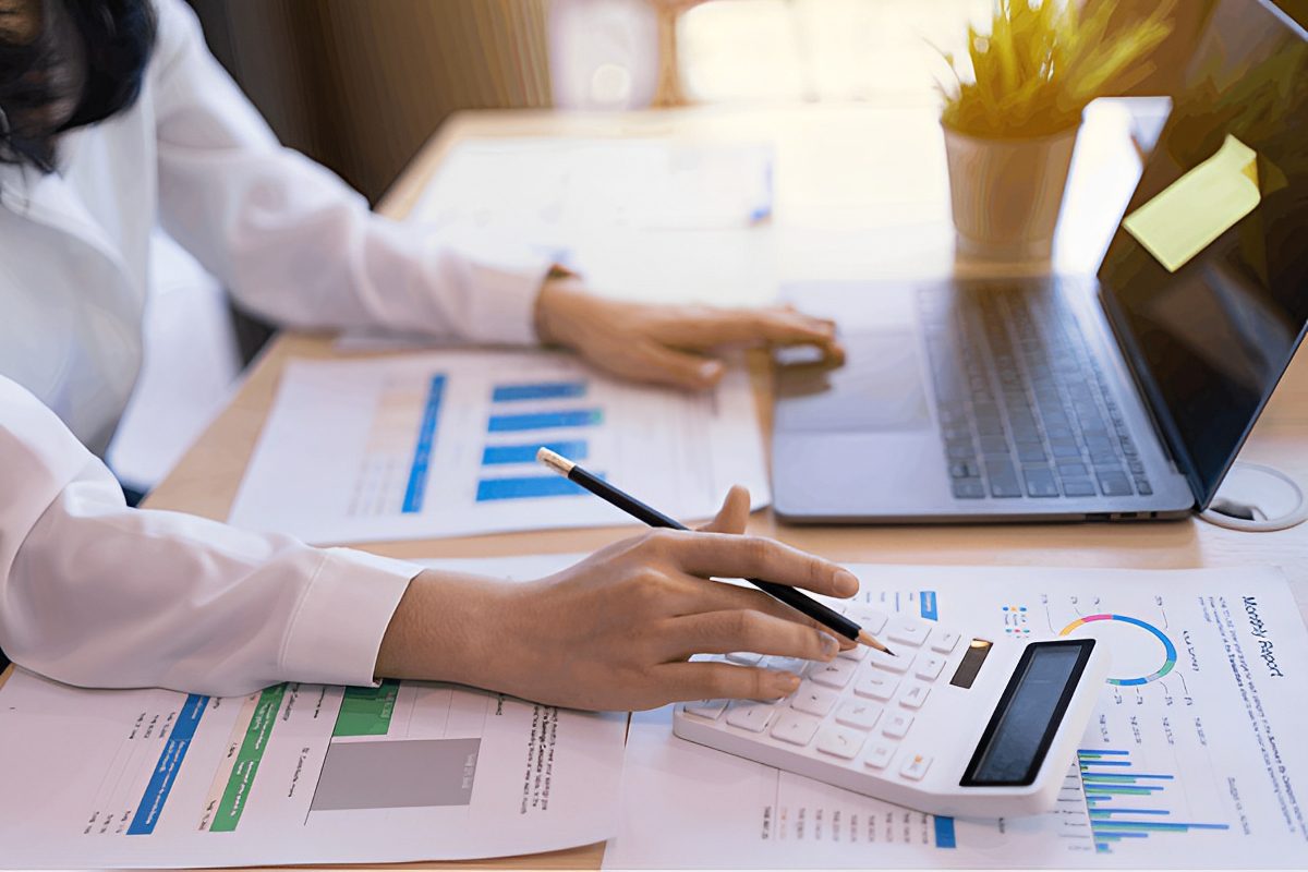 A person uses a calculator while analyzing financial graphs and charts on a desk with a laptop and plant nearby.