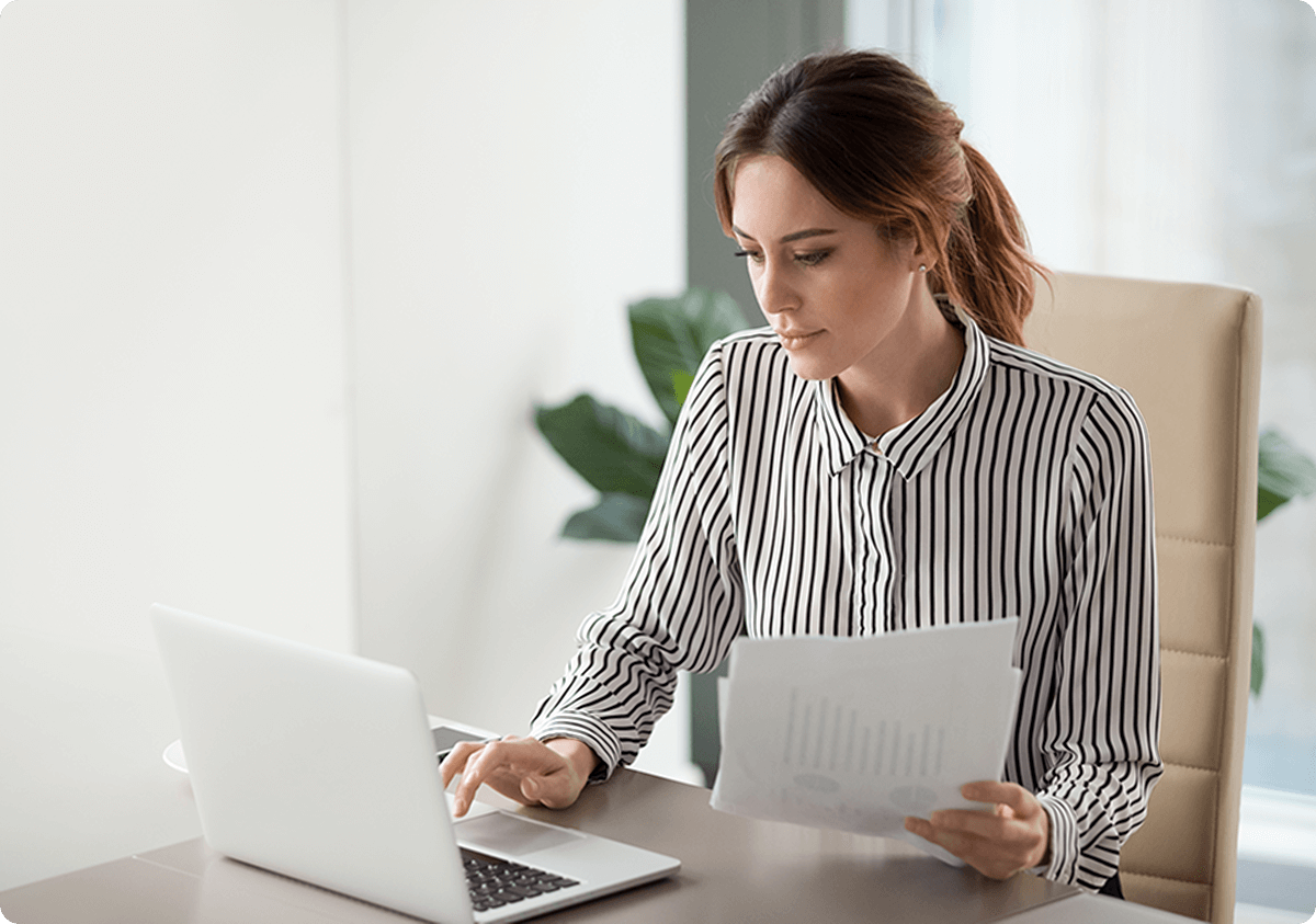 A woman in a striped shirt sits at a desk, working on a laptop while holding papers.